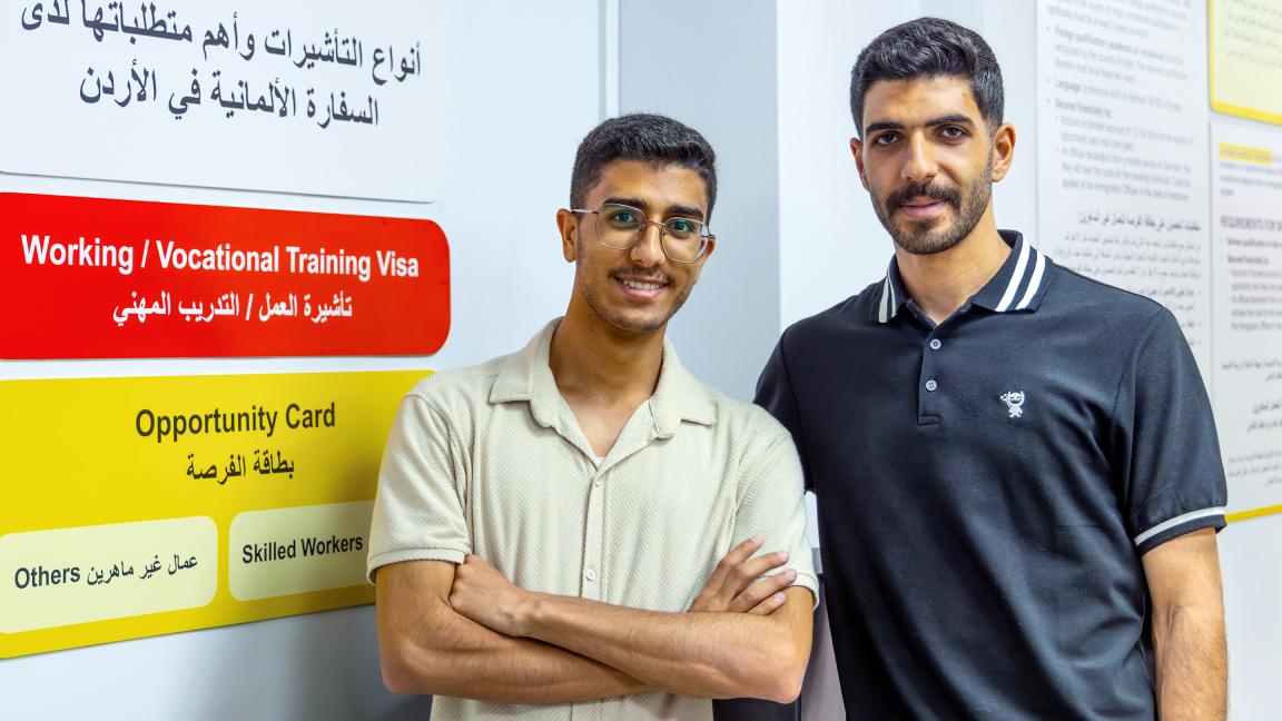Two young men stand side by side in front of an informational poster about visa and work opportunities in Jordan.