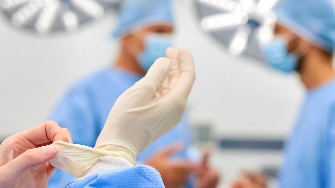 A medical professional is putting on sterile gloves in an operating room, with two other people in surgical attire blurred in the background.