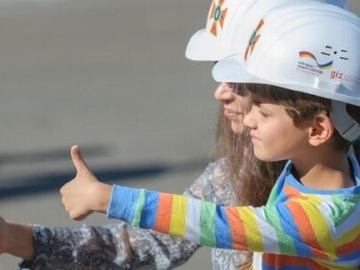 Frau hält einen Jungen im Arm. Beide haben einen weißen Helm auf und zeigen den Daumen nach oben