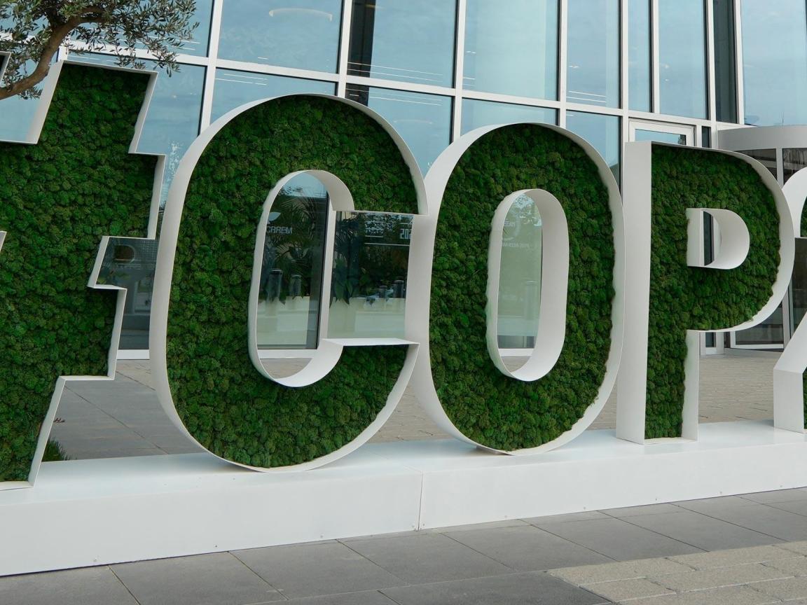 The image shows a large ‘COP29’ sign, whose letters are filled with green plants, in front of a modern glass building.