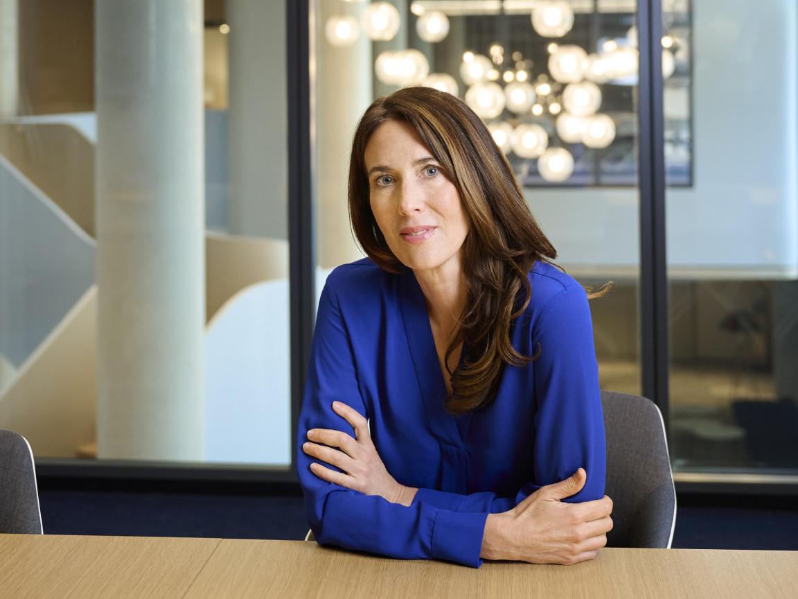A woman with long brown hair wearing a blue blouse sits at a conference table in a modern office with glass walls and round ceiling lights in the background.