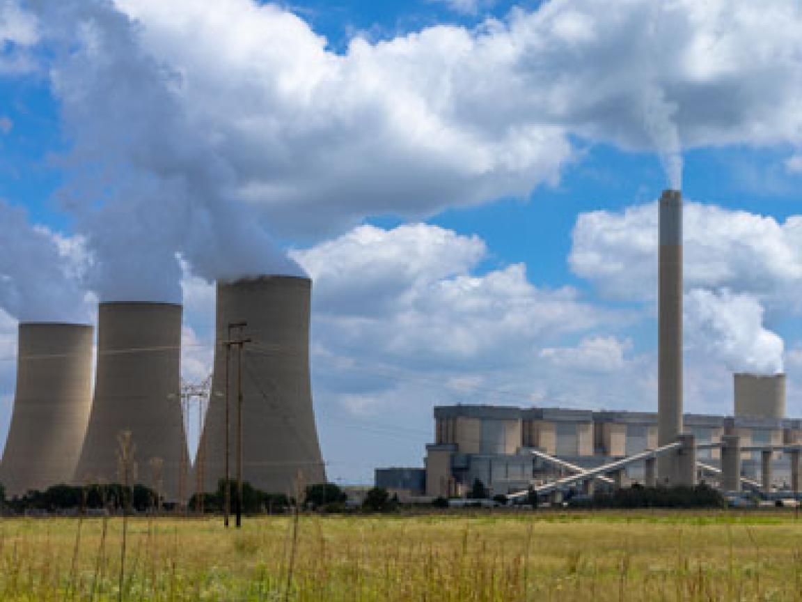Ein weites Feld im Vordergrund mit einem Kraftwerk im Hintergrund unter einem teilweise bewölkten Himmel. Das Kraftwerk hat mehrere hohe, schlanke Schornsteine und Kühltürme, aus denen Dampf aufsteigt
