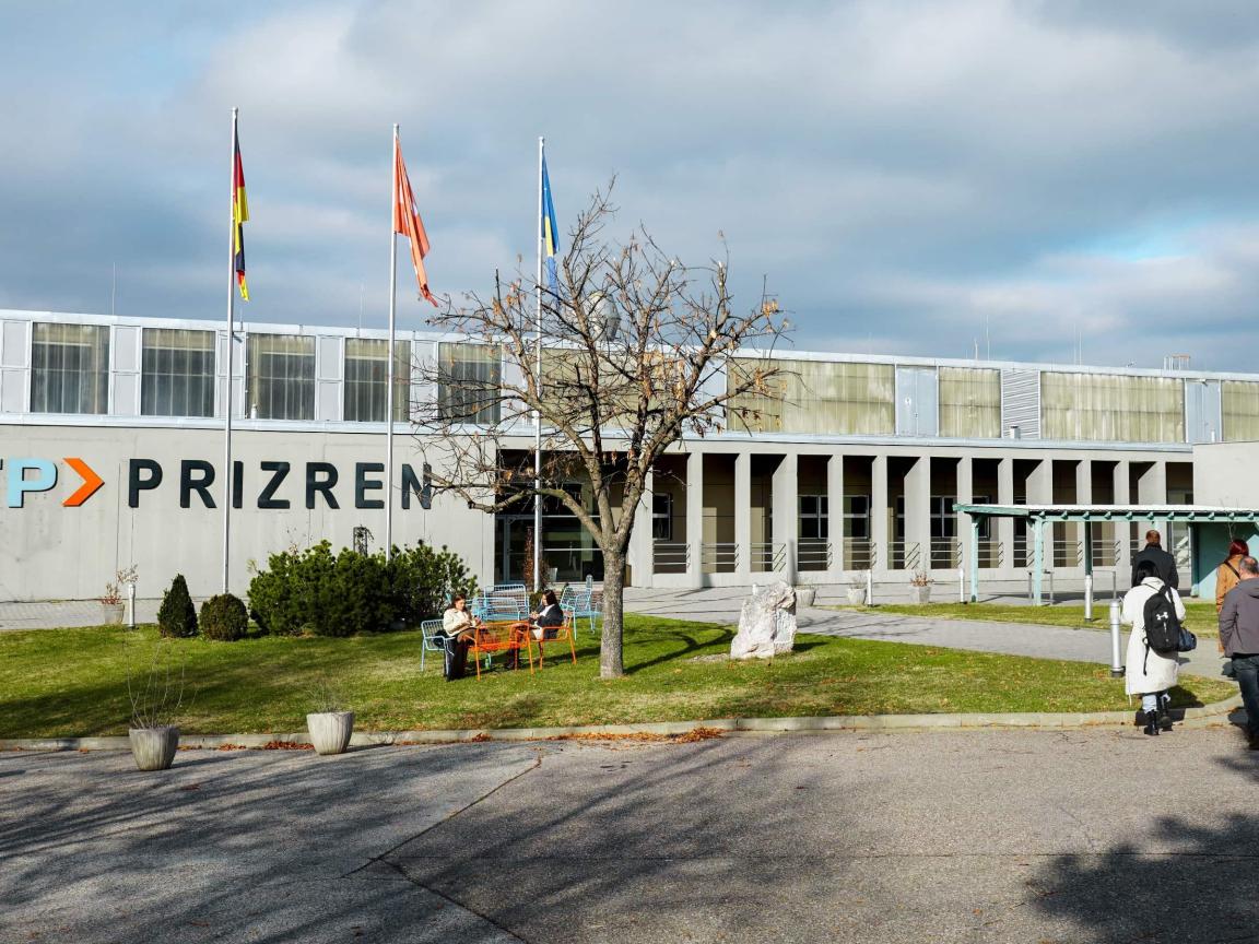 Exterior view of the ITP Prizren building with three flagpoles, seating on the lawn and several people on the grounds.
