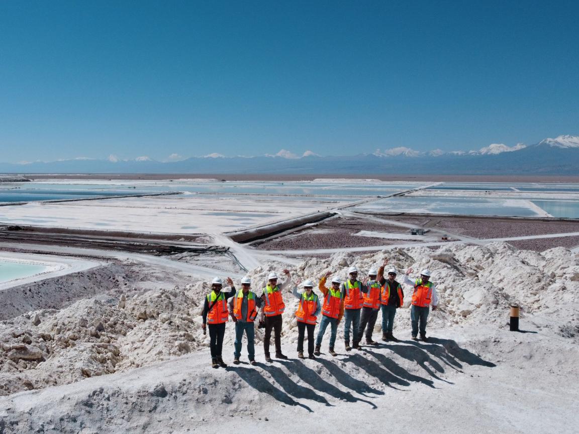 Gruppe von Personen mit Sicherheitshelmen und leuchtend orangen Warnwesten steht auf einem Hügel in einem Salzabbaugebiet mit grossen Verdunstungsbecken; im Hintergrund sind Berge und ein klarer blauer Himmel sichtbar.