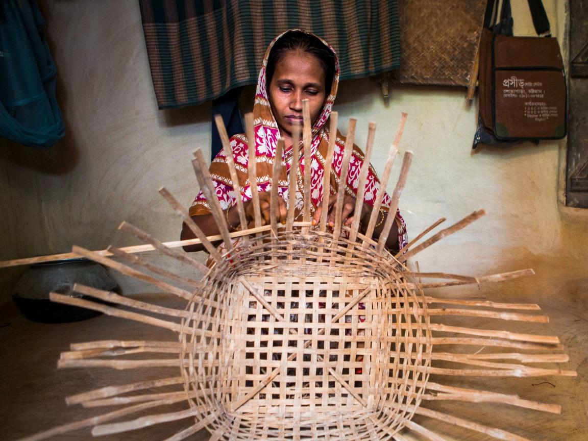 Eine Frau in traditioneller Kleidung sitzt in einem einfachen Innenraum und flechtet konzentriert einen grossen Korb aus Bambus oder ähnlichem Naturmaterial.       ChatGPT fragen