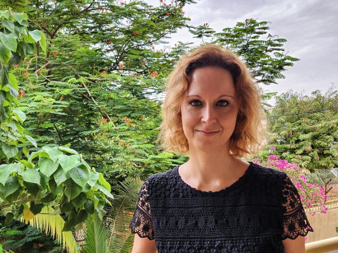 Eine Frau mit blonden, lockigen Haaren steht auf einem Balkon, trägt ein schwarzes, gemustertes Spitzenoberteil und lächelt leicht, im Hintergrund ist üppiges tropisches Grün mit Blüten und einem bewölkten Himmel zu sehen.
