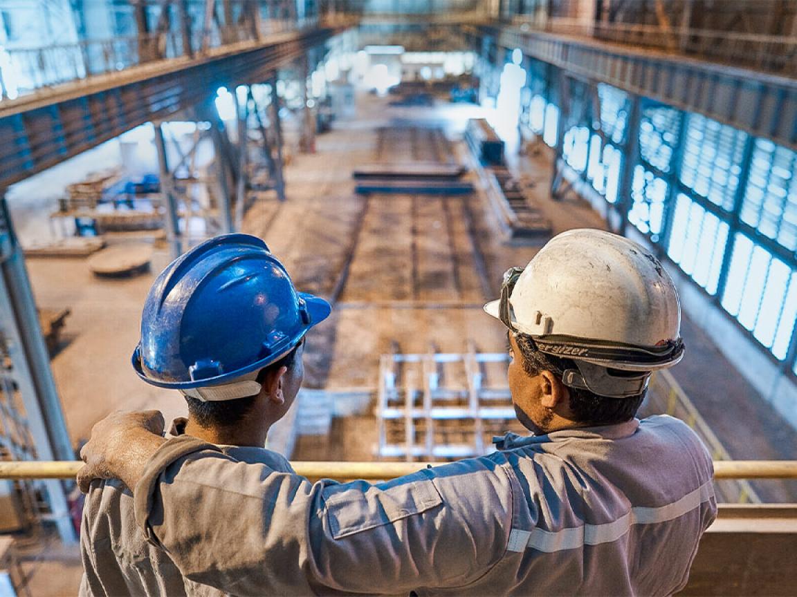 Two men in hard hats stand on a gallery looking down on a large industrial hall with machines and work areas.