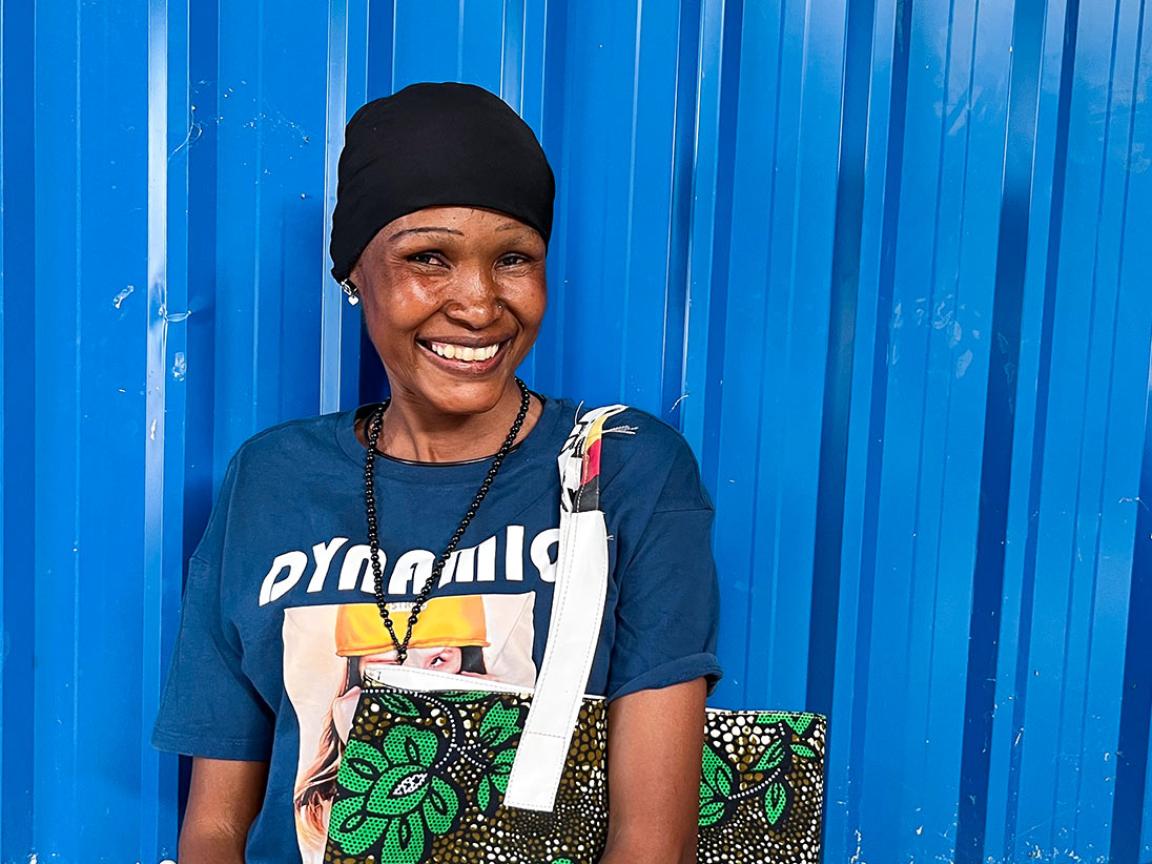 Eine lächelnde Frau steht vor einer blauen Metallwand, trägt ein bedrucktes T-Shirt und eine bunte Stofftasche mit Blumenmuster.