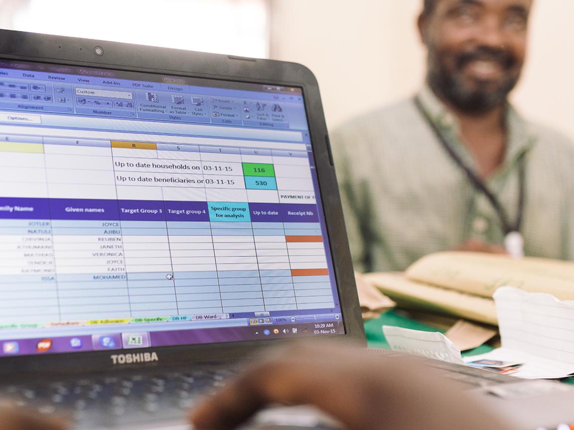 Ein Mensch tippt auf einem Laptop mit Excel-Tabelle, im Hintergrund sitzt ein lächelnder Mann an einem Schreibtisch.