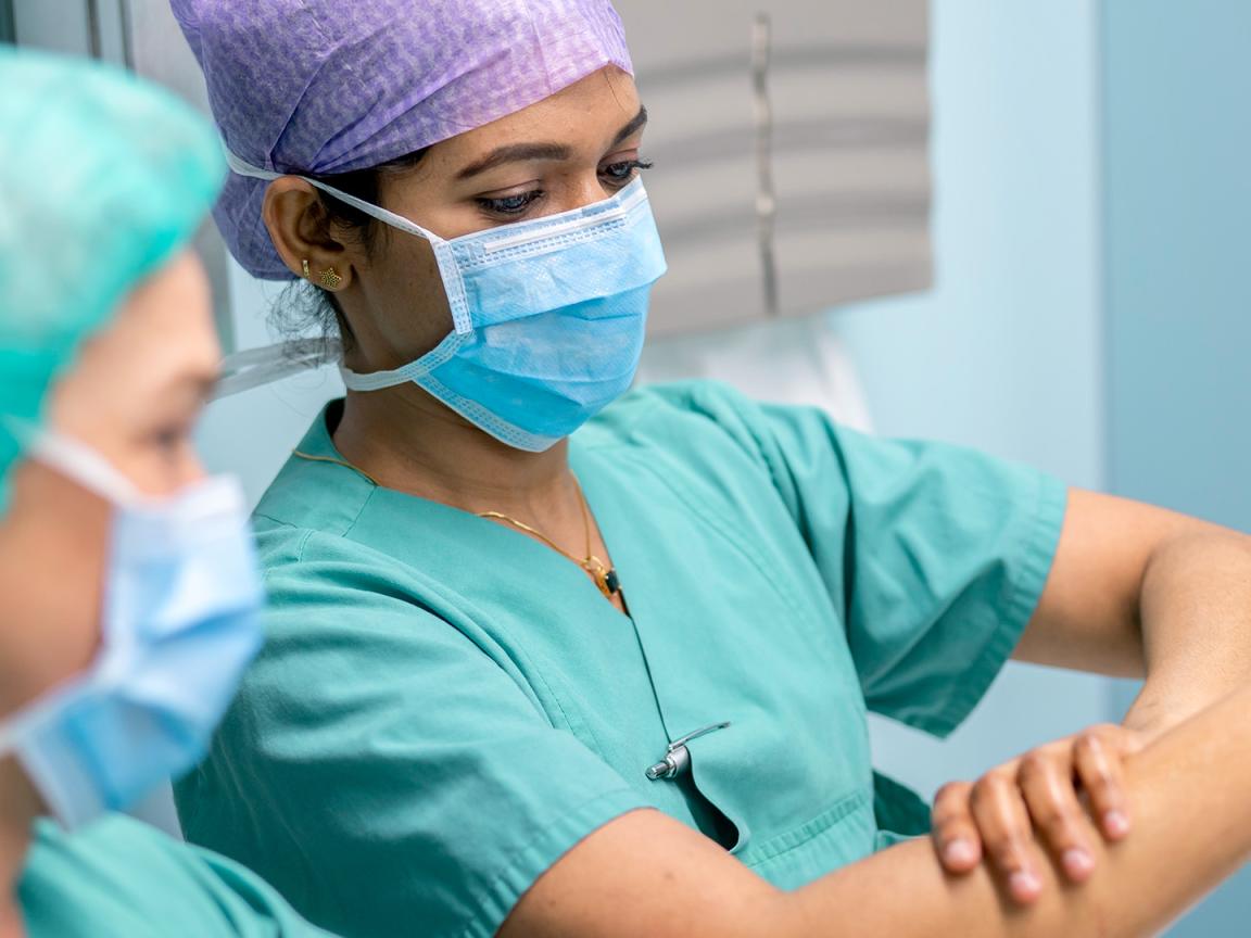 Two surgical staff members in green scrubs and face masks carefully disinfect their forearms.