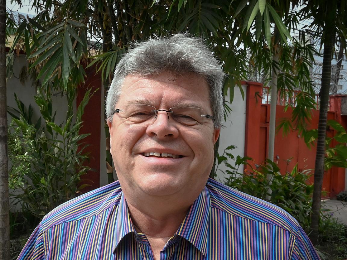 A man with gray hair and glasses smiles at the camera, wearing a brightly striped shirt and standing outdoors in front of tropical plants.