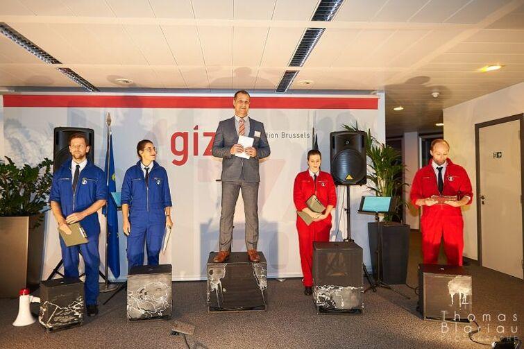 In der Mitte steht ein Mann auf einem Podest. Links stehen zwei Personen in blauen Overalls und rechts zwei Personen in roten Overalls