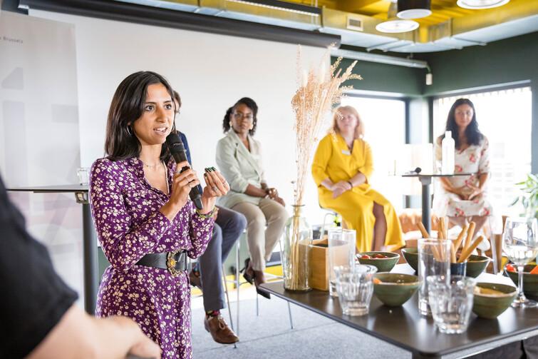 Frau steht mit einem Mikrofon in der Hand vor 4 sitzenden Personen