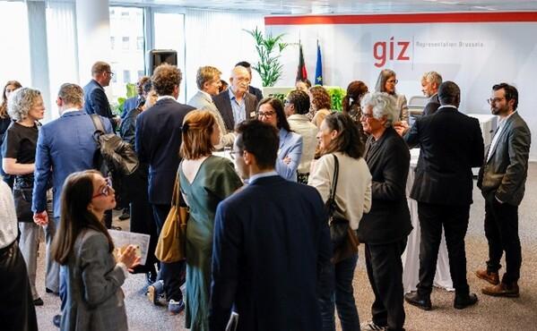 Viele Menschen stehen verteilt in einem Raum. Hinten an der rechten Wand steht "GIZ Representation Brussels"