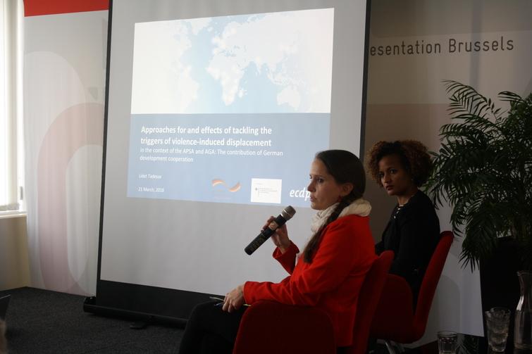 Zwei Frauen sitzen seitlich vor einer Leinwand. Eine Frau hält ein Mikrofon