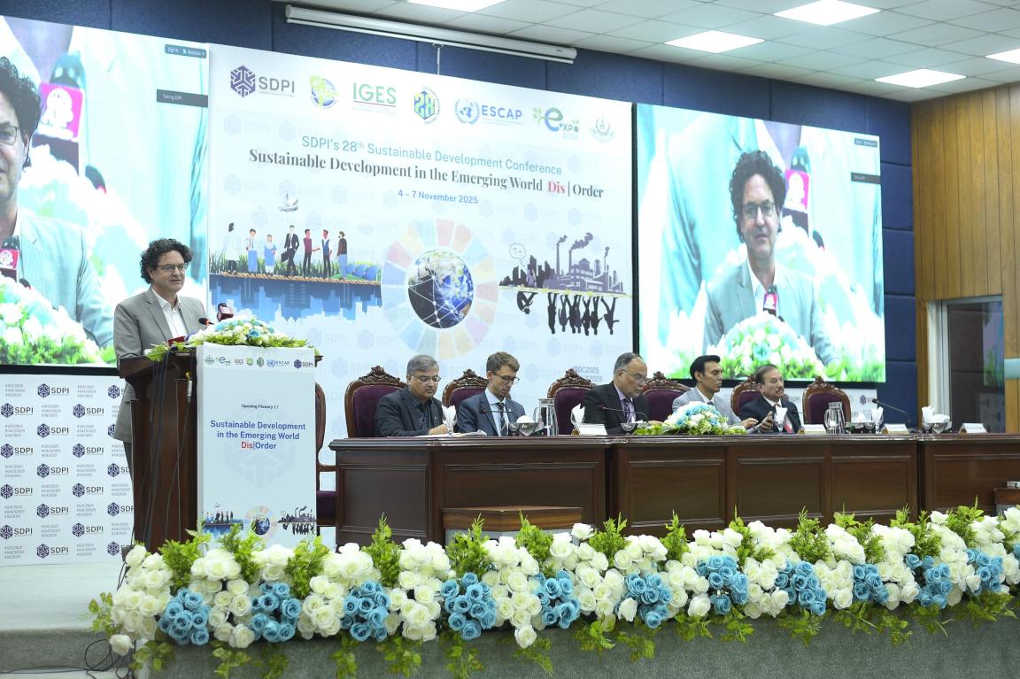 Mr. Arno Kirchhof speaking at the conference, with panelists seated and a large screen displaying the event title.