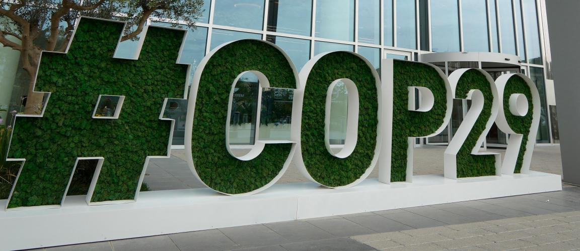 The image shows a large ‘COP29’ sign, whose letters are filled with green plants, in front of a modern glass building.