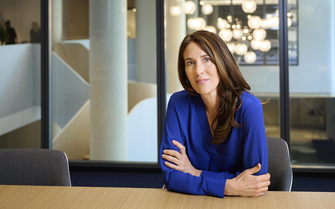 A woman with long brown hair wearing a blue blouse sits at a conference table in a modern office with glass walls and round ceiling lights in the background.