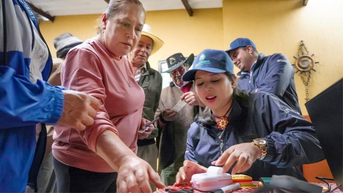 Eine Gruppe von Menschen versammelt sich um einen Tisch, wobei eine Frau im rosa Pullover zuschaut, während eine andere mit blauer Mütze ein kleines Gerät bedient.