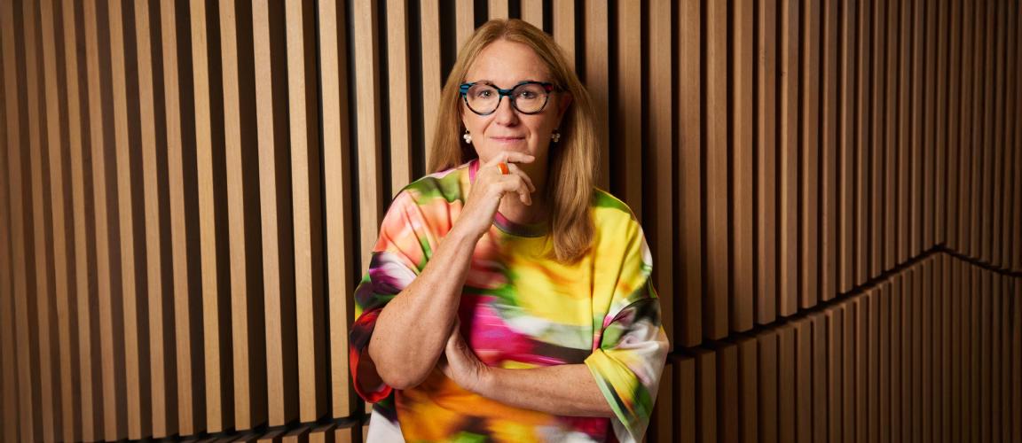 A woman with long blonde hair and glasses stands in front of a wall with wooden slats and wears a colourful blouse.