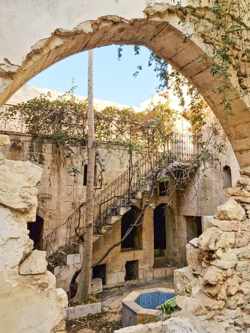 Dilapidated courtyard with stone arch, weathered stone steps and a fountain, partially overgrown with plants.