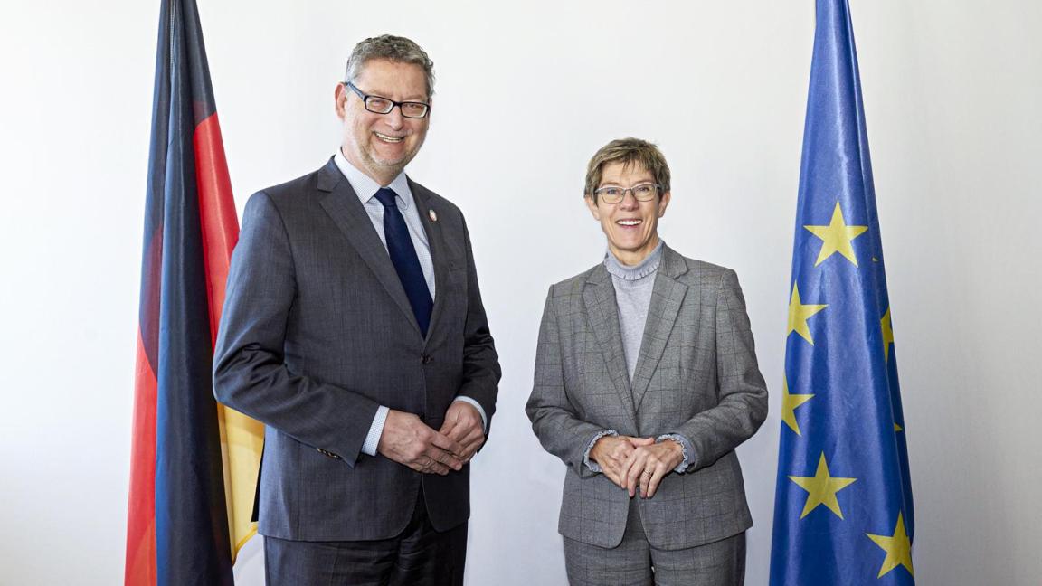 Two people in formal attire stand side by side and smile at the camera, flanked by a German flag on the left and a European flag on the right against a light background.
