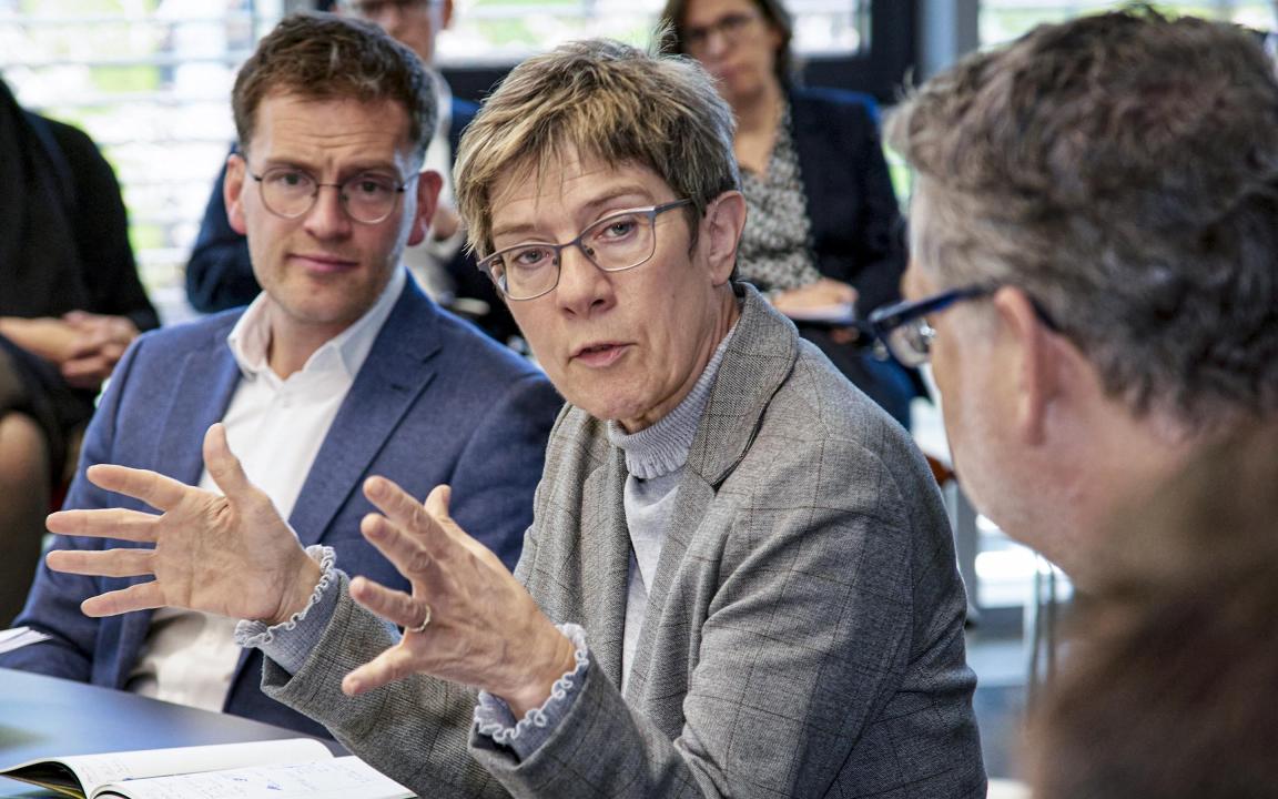 Discussion in a conference room with a woman in a grey blazer speaking animatedly, surrounded by several people in business attire listening attentively.