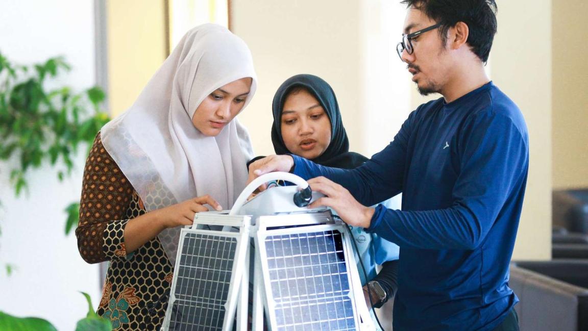Zwei junge Frauen mit Hijab und ein Mann untersuchen gemeinsam ein technisches Gerät mit Solarpanels in einem Innenraum.