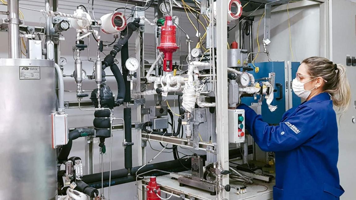 A woman wearing a blue lab coat and a face mask is working on a complex industrial apparatus with numerous pipes, gauges and valves.