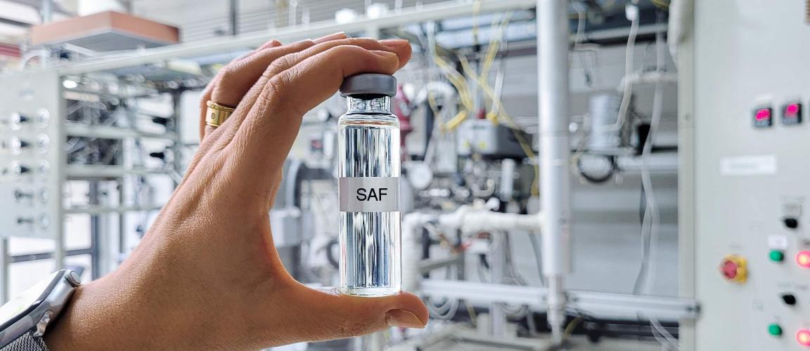A hand holds a small bottle labelled ‘SAF’ containing a clear liquid against a blurred industrial background.