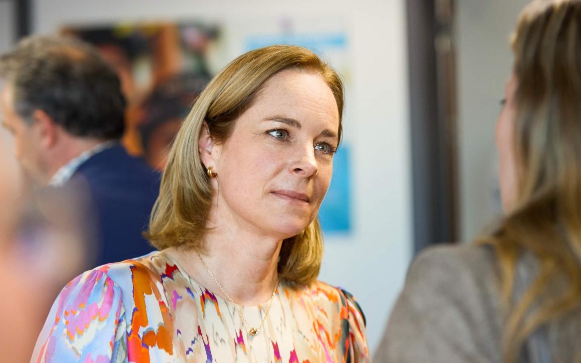 A woman with shoulder-length blonde hair and a colourful blouse talking to another person, with blurred people and pictures on the wall in the background.