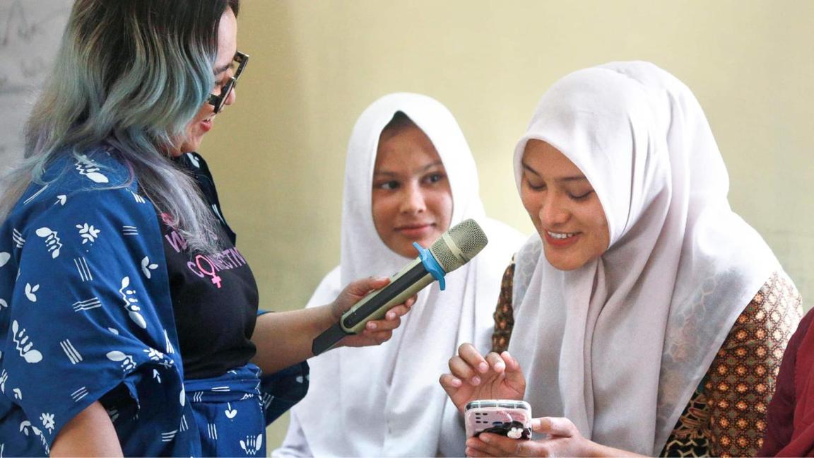 A woman with a microphone interviews two young women wearing headscarves, one of whom is using a smartphone.