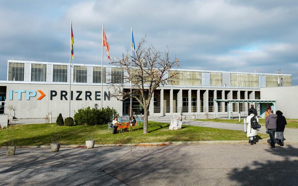 Exterior view of the ITP Prizren building with three flagpoles, seating on the lawn and several people on the grounds.