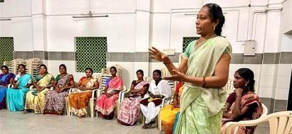 A woman stands in a circle of seated textile workers and explains something.Textile workers work in a factory.