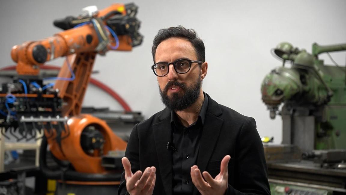 A man with glasses and a dark beard gesticulates in an industrial setting with an orange-coloured robot arm and a green machine in the background.