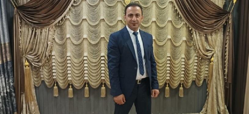 Ein Mann in einem Anzug mit Krawatte steht vor einer kunstvoll gerafften Vorhanginstallation in Gold-, Beige- und Brauntönen.