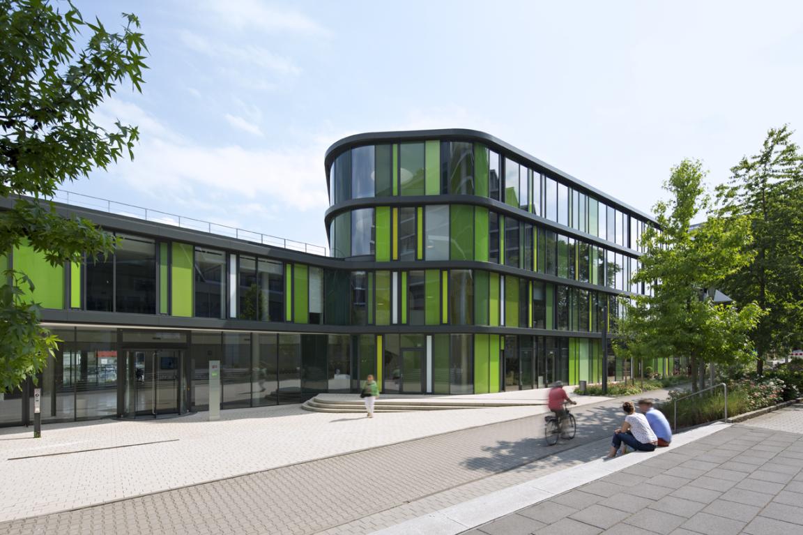 Ein modernes, organisch geformtes Gebäude mit vielen Fenstern und hellgrünen Flächen. Im Vordergrund sitzen zwei Personen auf einer Treppe, ein Radfahrer fährt vorbei.