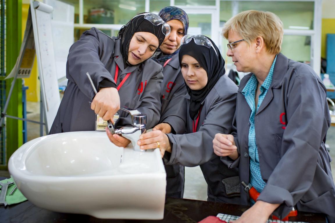 Foto: Vier Frauen in Arbeitskitteln stehen um ein Waschbecken. Eine Frau schraubt am Wasserhahn, eine weitere Frau hält ihn.