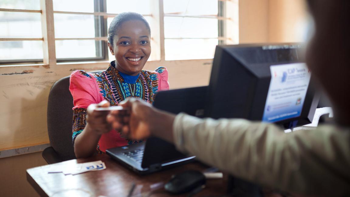 Smiling woman in colorful traditional clothing sits at a desk with a computer and hands a card or document to a person opposite her.