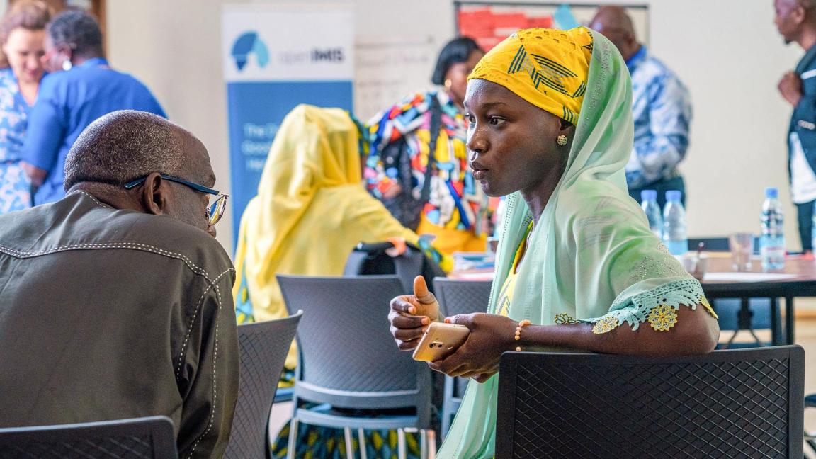 Frau in gelbem Kopftuch und hellgrüner Kleidung spricht in einem Konferenzraum ernsthaft mit einem älteren Mann; im Hintergrund sind weitere Teilnehmende und Infotafeln sichtbar.