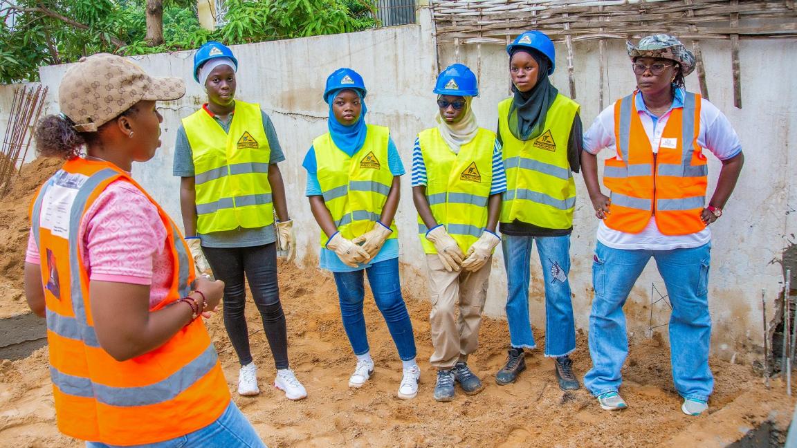 Group photo with Djélia Diagne (left, in profile) talking to five young women wearing protective helmets and high-visibility vests on a construction site.