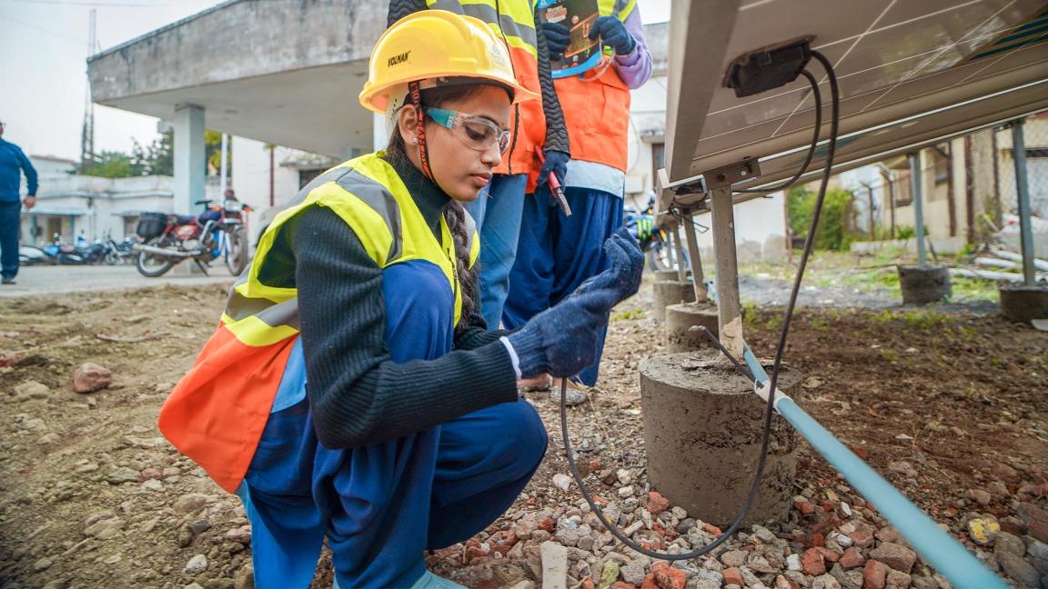 Eine junge Frau in Schutzkleidung überprüft Kabel unter einem Solarpanel, zwei weitere Personen stehen mit Werkzeug und Unterlagen daneben.