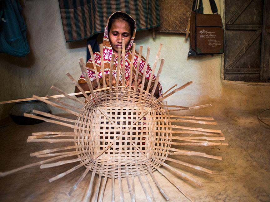 A woman sitting in a room hand-weaves a large basket made of wooden strips.