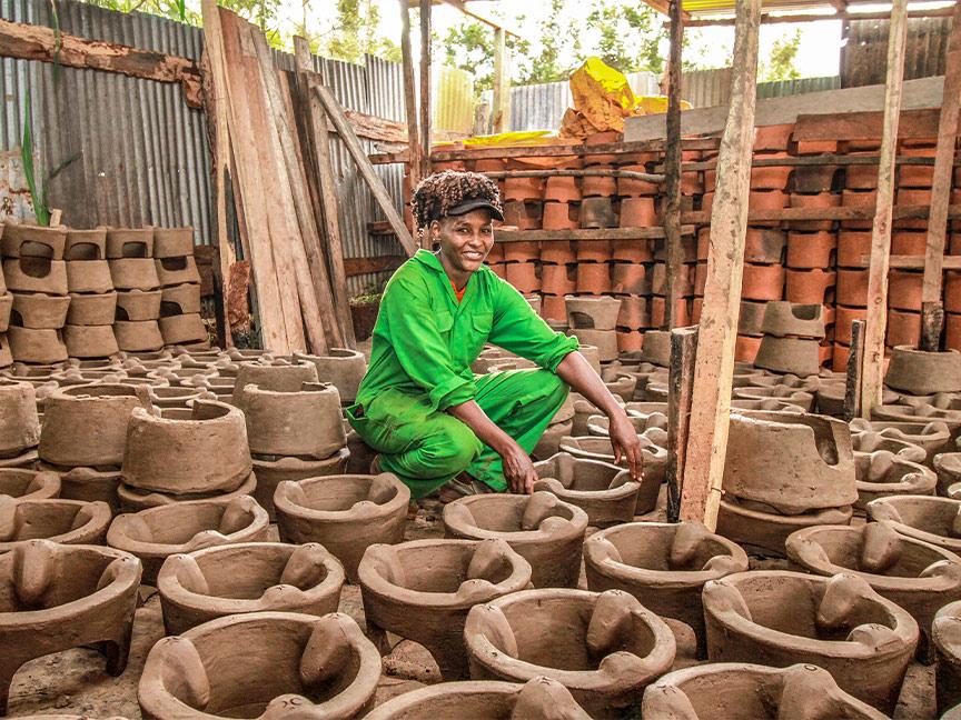 A woman wearing green overalls smiles and crouches among many clay ovens in a workshop.