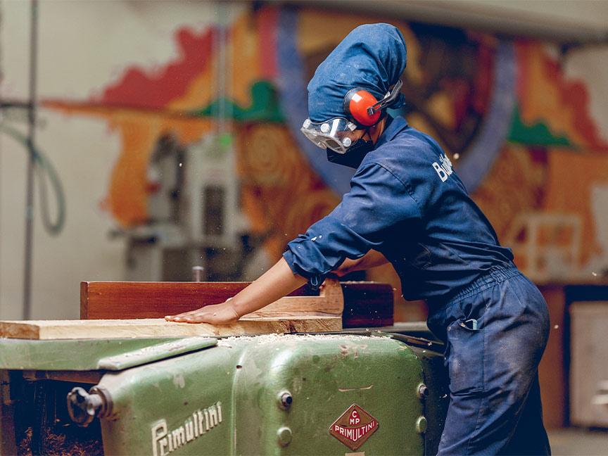 A woman in overalls and protective gear uses a woodworking machine in a workshop with a colourful wall mural.