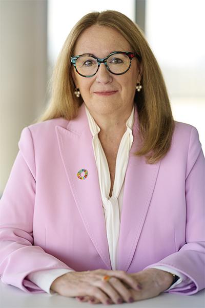 A woman with long blonde hair wearing a pink blazer and colorful glasses sits against a bright background, wearing an SDG color wheel pin.