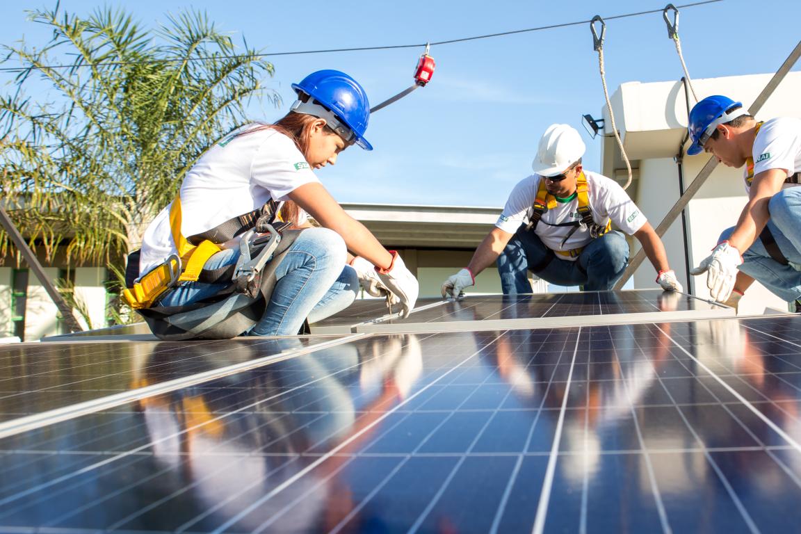 Three people wearing helmets are installing solar panels and are secured by a rope.