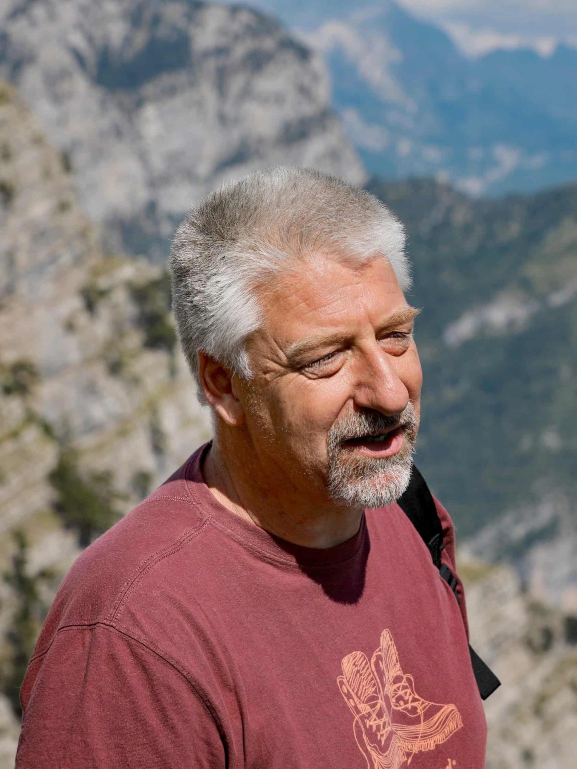 An older man with gray hair and beard stands in the mountains wearing a maroon T-shirt with a hiking boot graphic.