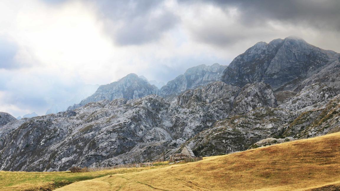 A small wooden hut stands on a meadow in front of steep, rocky mountain peaks under a dramatic cloudy sky.