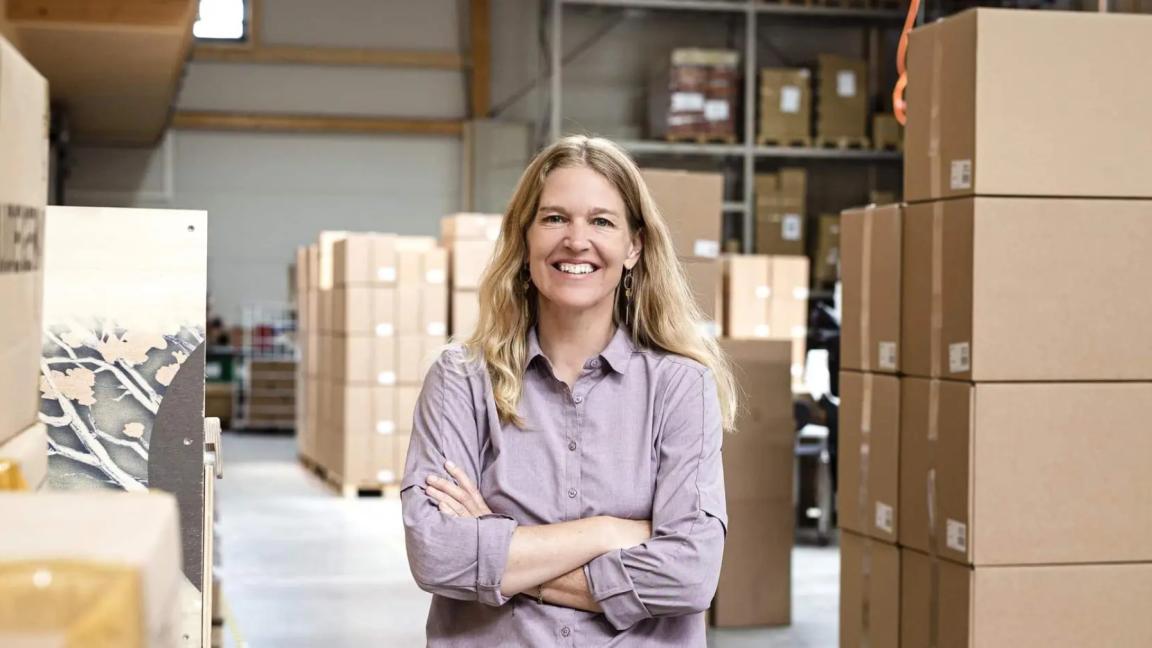 A smiling woman with long blond hair stands with crossed arms in a warehouse full of cardboard boxes.
